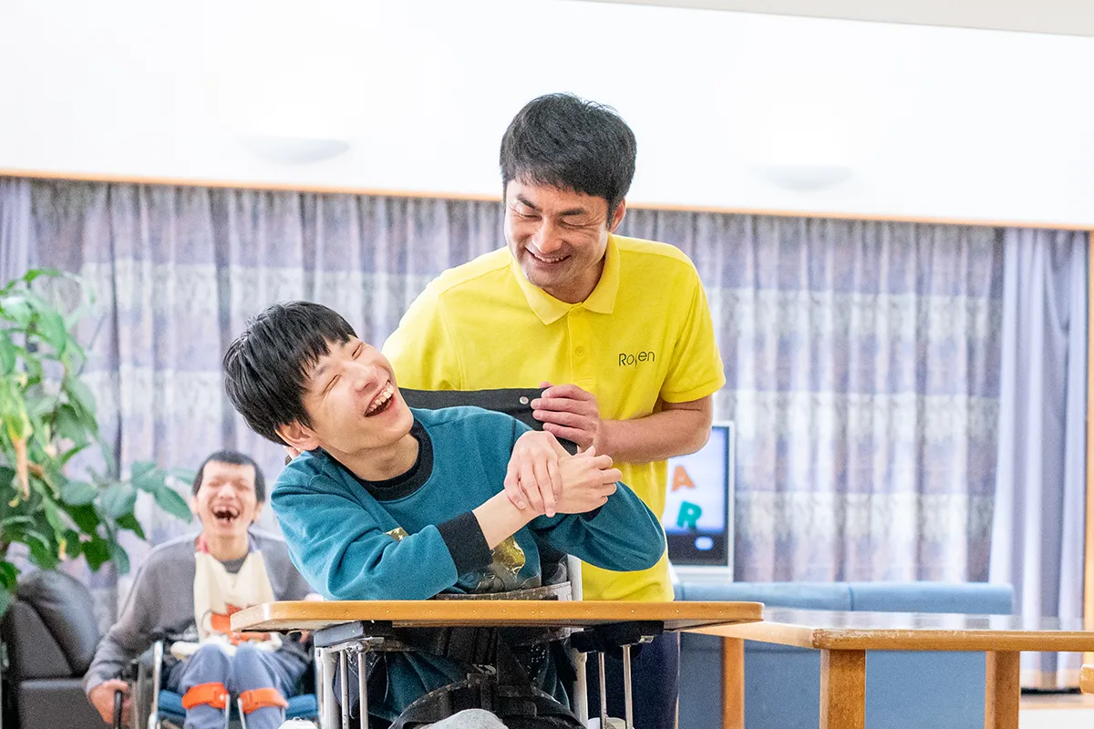 介護職員の男性が、利用者の男性の椅子を支えている様子